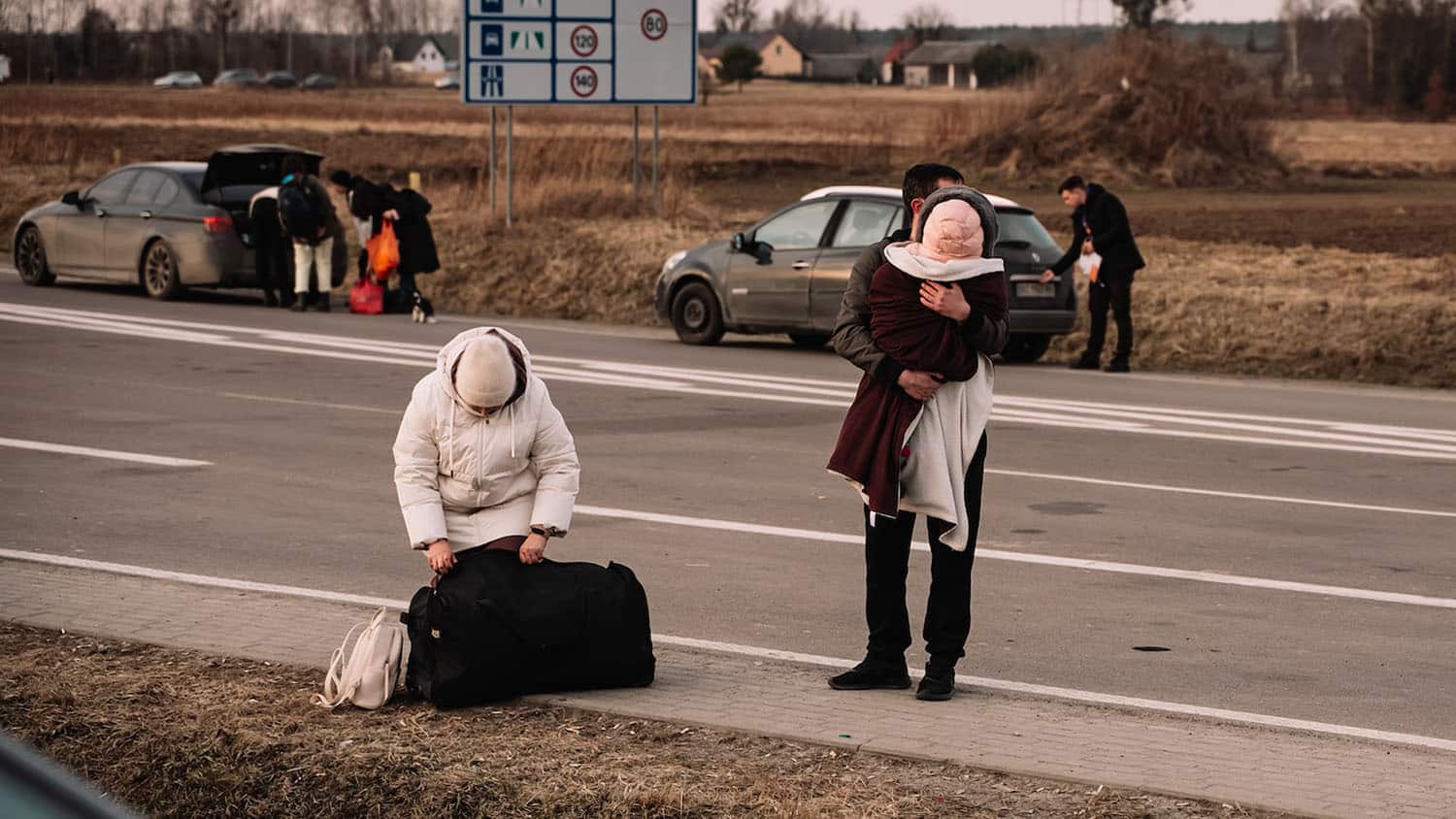 两名成年乌克兰难民站在路边。其中一个抱着一个孩子。路牌告诉你这是在欧盟境内。