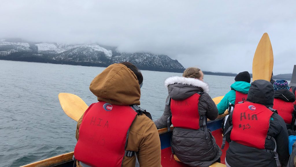 学生们穿着厚重的外套和救生衣，划着独木舟穿过阿拉斯加海岸，远处是雪山