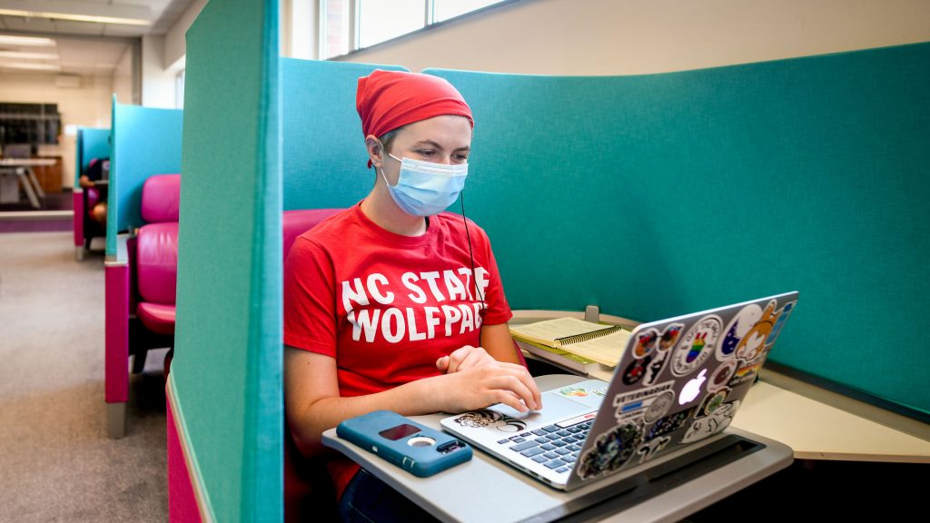 一名戴着面罩和一个NC状态T恤学习的学生在山图书馆的一把蓝色椅子上。