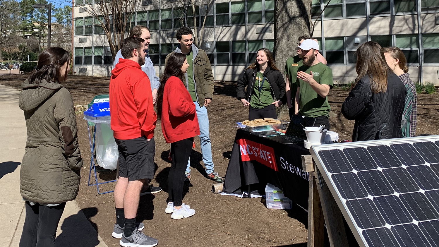 北卡罗来纳州立大学的管理人员穿着配套的绿色t恤，在室外的一块太阳能电池板旁边对一小群同学讲话。