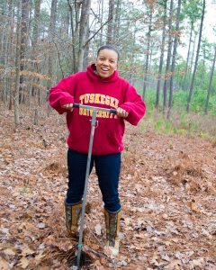 Zakiya Leggett  - 在一个红色的tuskegee大学连帽衫，牛仔裤和棕色靴子 - 在树木繁茂的地区外面采取土壤样本。