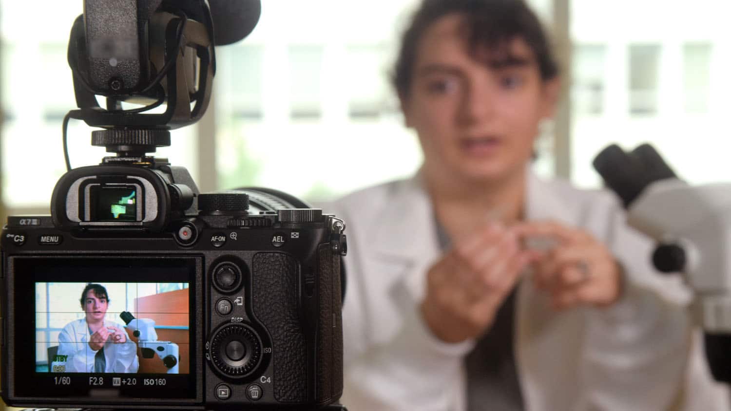 一台照相机正在拍摄一位科学家谈论她的工作;在相机的取景器中也可以看到这位科学家
