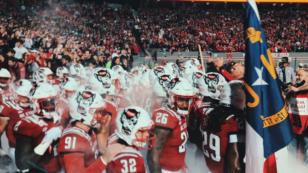 The NC State basketball team standing on the sidelines, with a packed stadium behind them.