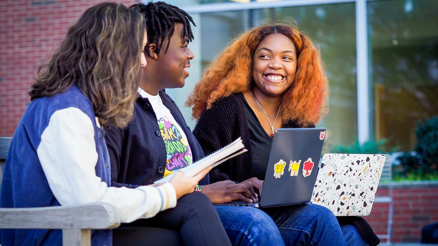 Three students sit on a bench with their laptops on their laps.