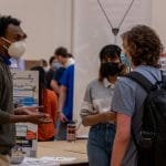 Three masked students talk near a poster at the Winter Involvement Fair.