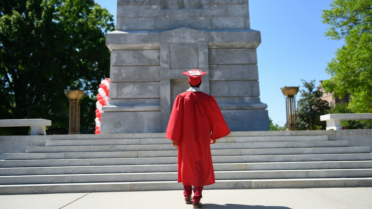 Graduating student dressed in red regalia faces NC State Belltower