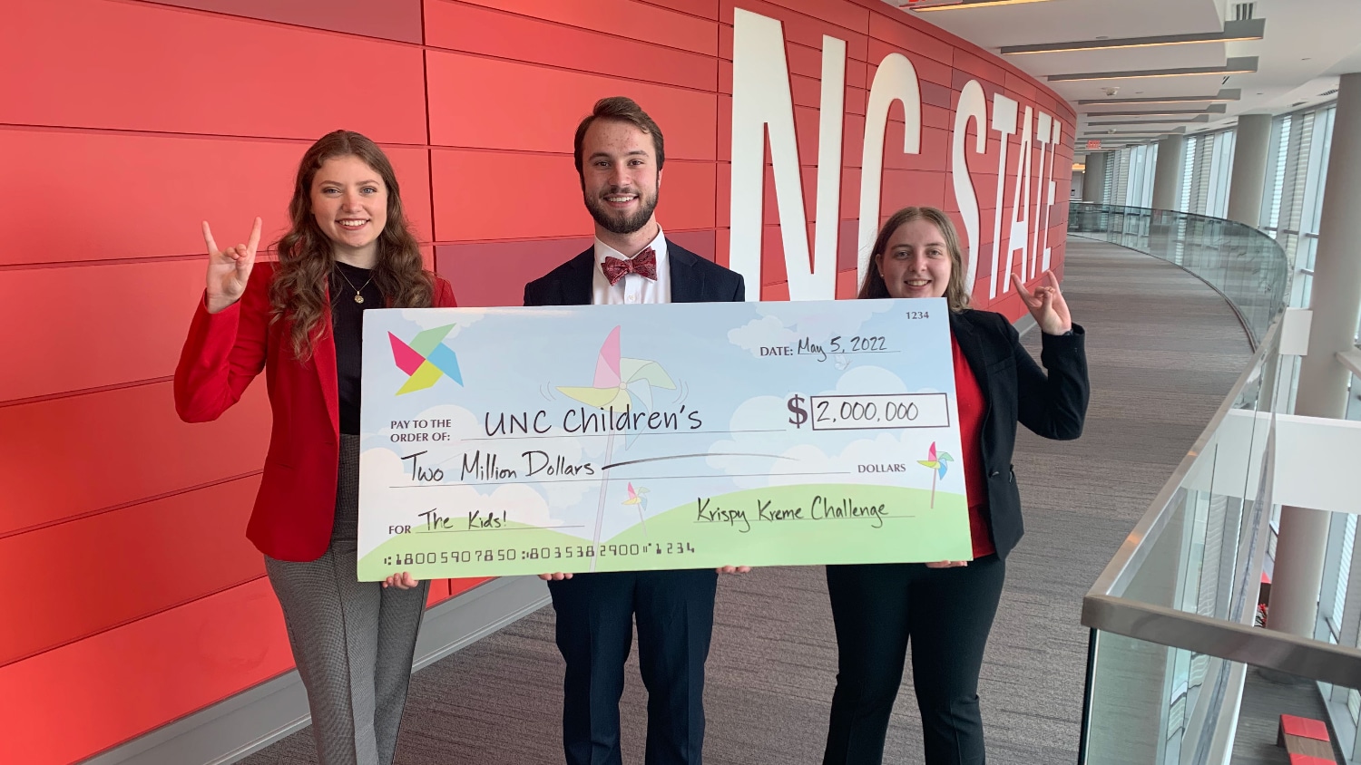 2022 Krispy Kreme Challenge Race Directors pose with $2M commitment check for UNC Children's and hold the Wolfpack sign.
