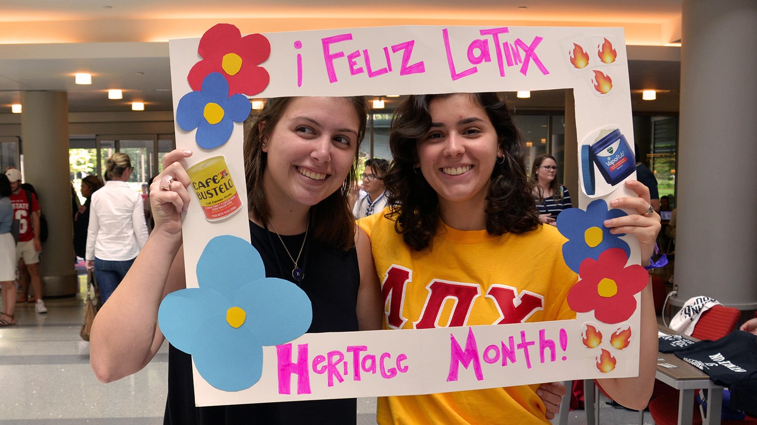 两名北卡罗来纳州立大学的学生在一个写着“Feliz latin Heritage Month”的纸板招牌的框架内微笑。