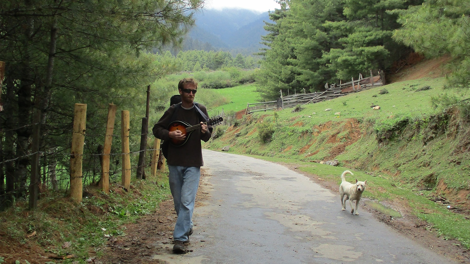 Ben Rose walks down a path in a wooded area while playing a mandolin. A white dog walks beside him.