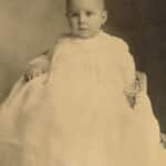 An infant Mary Yarbrough looks back at the camera in a sepia-toned portrait while wearing a large white dress.