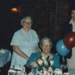 A blurred, color photo shows an adult Mary Yarbrough smiling at the camera while seated at a table. Several women, also smiling at the camera, sit and stand nearby