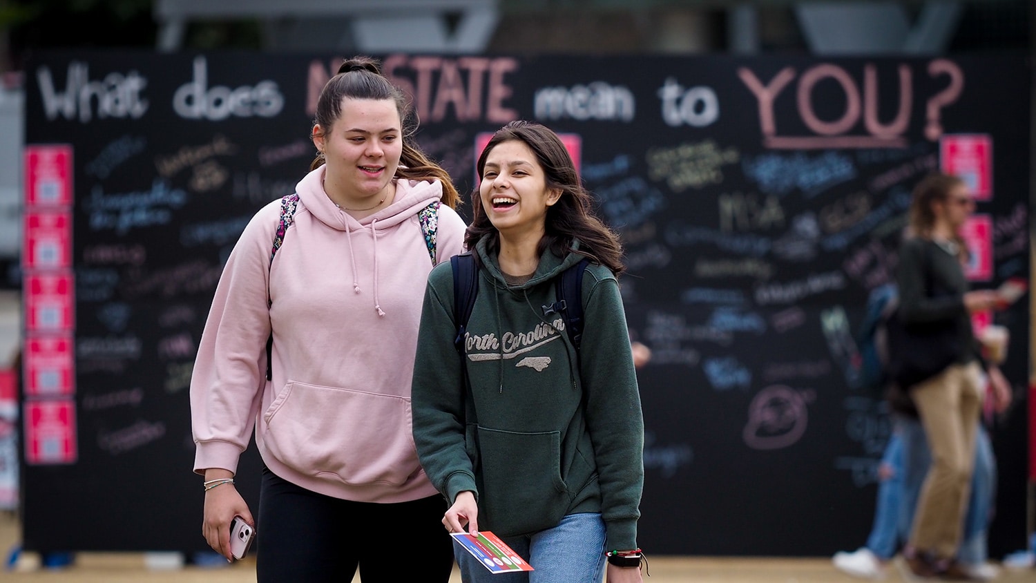 Two students laugh together while walking in front of a chalkboard sign that reads 