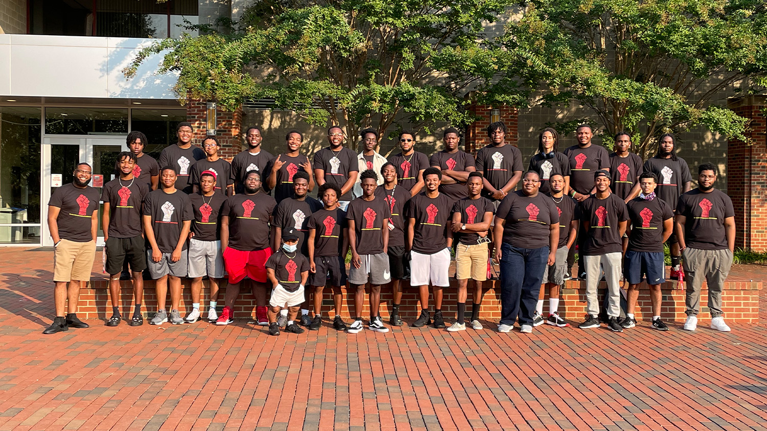 A large group of male students in black T-shirts stand on a brick path at NC State
