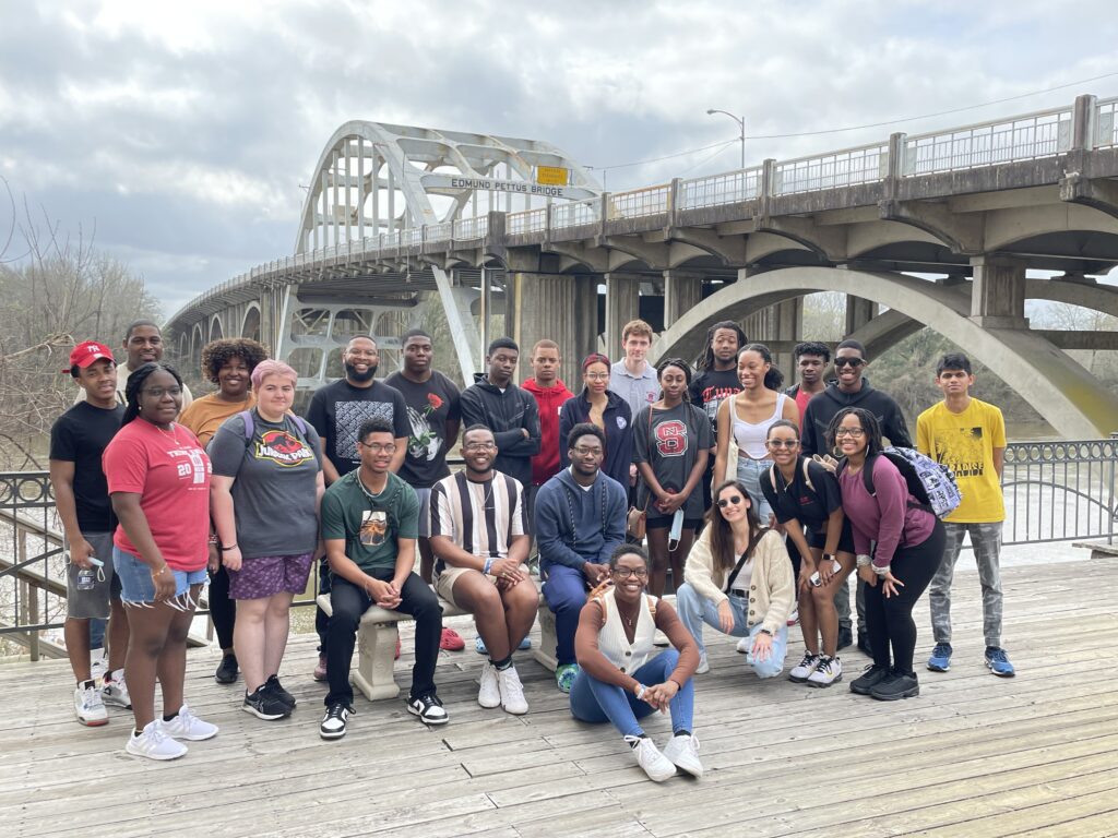 A large group of NC State students on a wooden board walk, with the Edmund Pettus Bridge in the background.