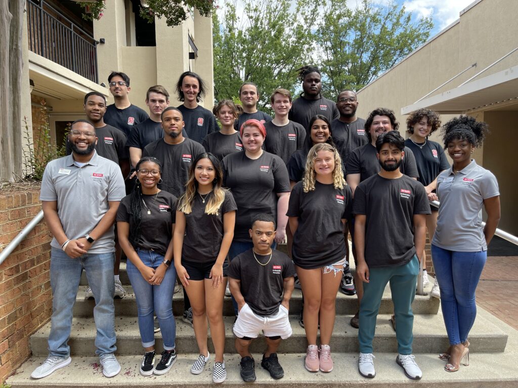 A large group of people in black University Housing T-shirts stands on outdoor stairs next to a residence hall