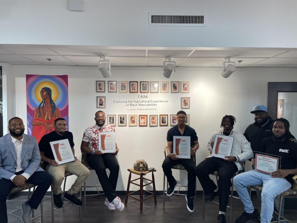 McKenzie and a group of students sit in a semi-circle and hold up their artwork in front of a wall decorated in photos.