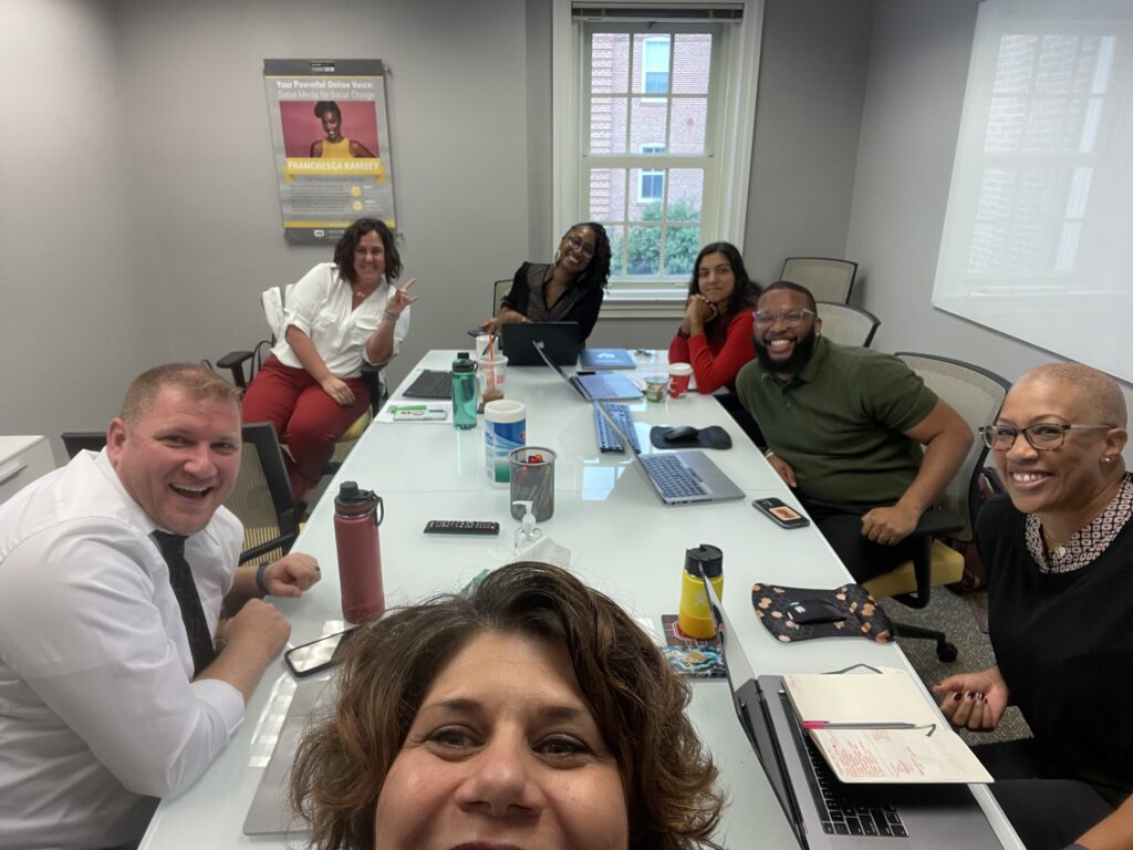 A selfie photo showing a group of people sitting around a white table in a conference room.