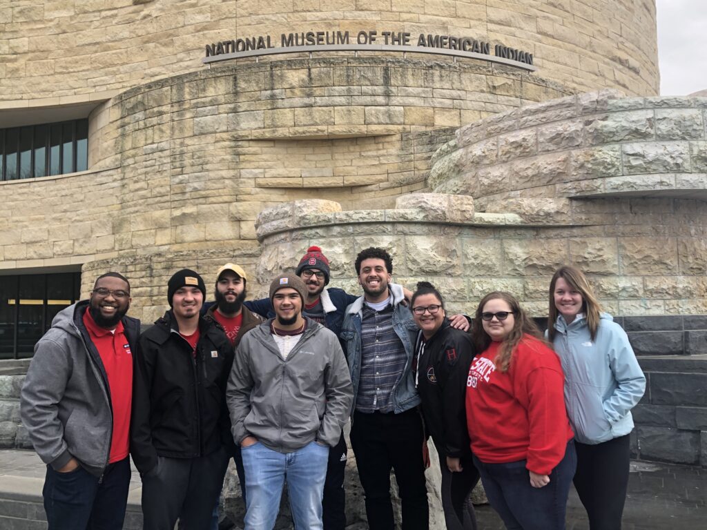 A group of people in red and black NC State clothing stand in front of a building with a sign that reads 