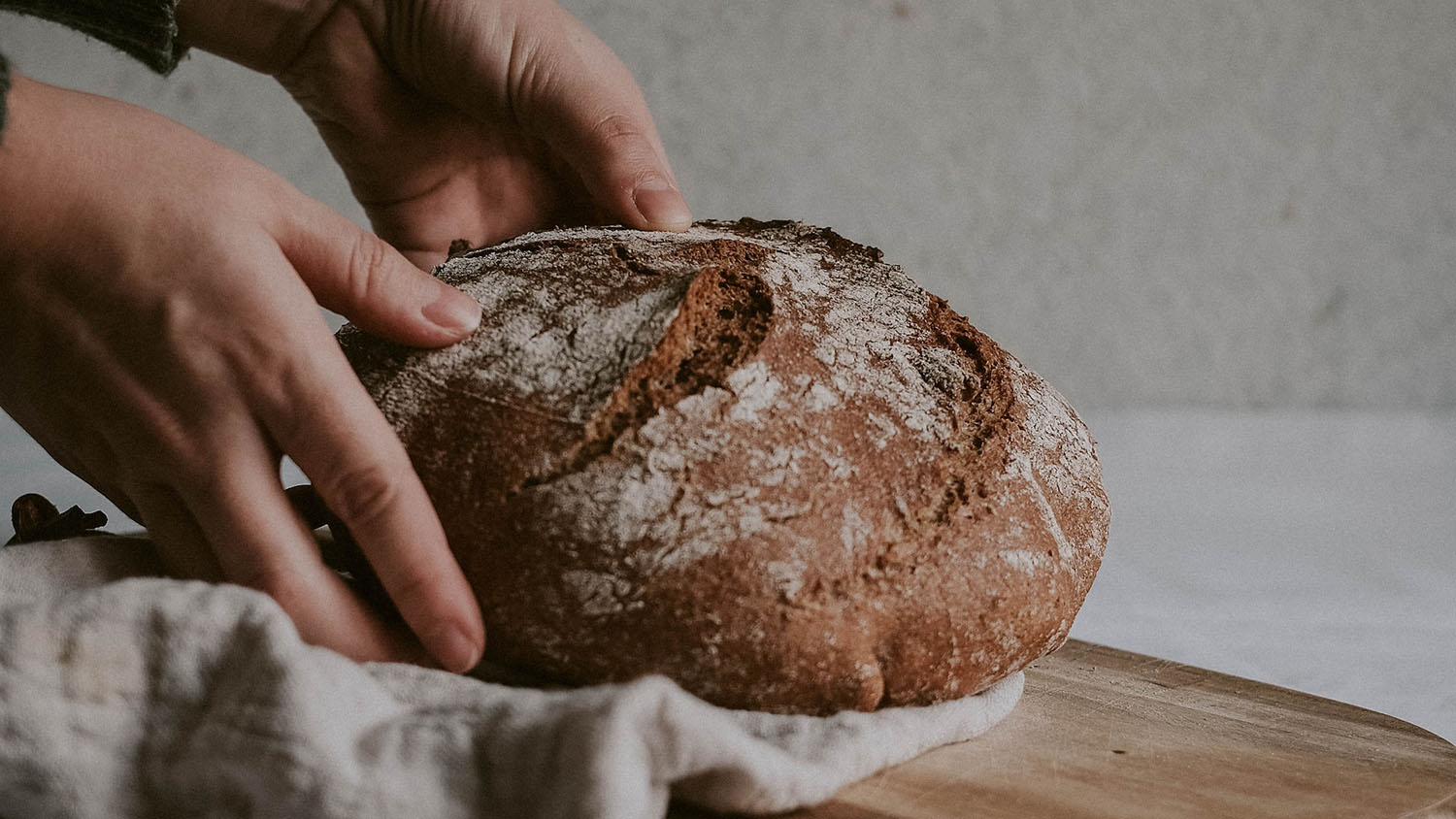 photo shows two hands placing a loaf of sourdough bread onto a wooden board draped with a cloth