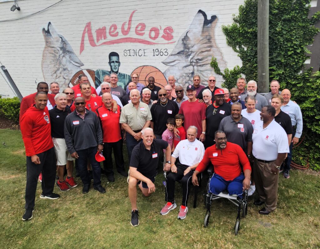 Bill Cowher stands among a large group of former NC State football players, coaches and his other friends from his time at NC State. Behind them is a white wall that reads 